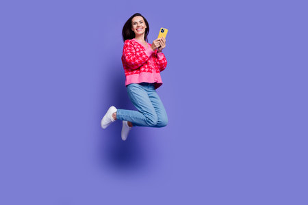 Full length photo of shiny attractive lady dressed pink cardigan jumping texting modern device isolated violet color backgroundの写真素材