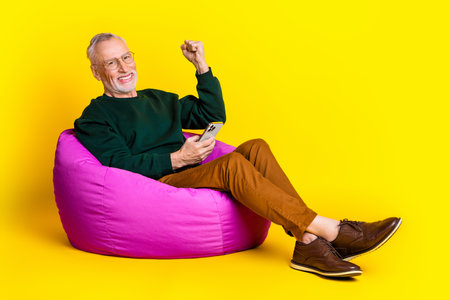 Full length photo of lovely pensioner man beanbag hold device winning dressed stylish green garment isolated on yellow color backgroundの写真素材