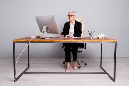 Full length photo of lovely pensioner lady startup boss sit table work computer dressed formalwear specs isolated on gray color backgroundの写真素材