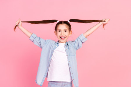 Photo of impressed small girl holding long ponytails dressed denim shirt having fun astonished staring isolated on pink color backgroundの写真素材