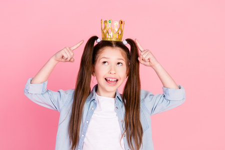 Photo of impressed cute small girl with ponytails dressed denim shirt look directing at crown on head isolated on pink color backgroundの写真素材