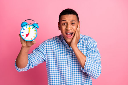 Photo of handsome impressed guy wear plaid shirt holding clock emtpy space isolated pink color...