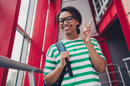 Photo of pretty young woman demonstrate v-sign striped t-shirt backpack work study business center officeの写真素材