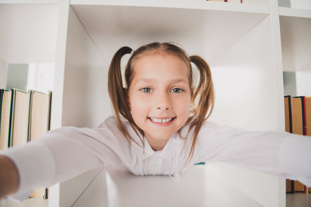 Photo portrait of cute adorable girl take book inside shelf library take selfie photo remote lesson education from home indoorsの写真素材