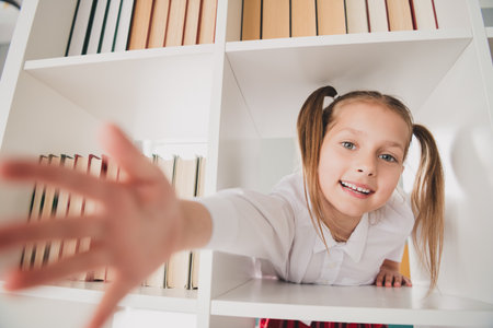 Photo portrait of small cute daughter stretch hand take book shelf library remote education home schooling isolated indoors backgroundの写真素材