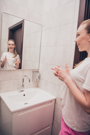 Vertical photo of pretty cheerful lady sleepwear pooking mirror pointing fingers herself indoors bathroom home houseの写真素材