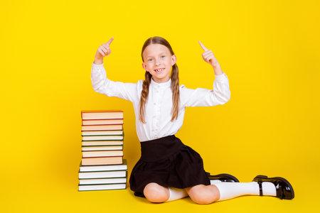 Full size photo of charming little pupil girl sit floor pile books point up empty space wear uniform isolated on yellow color backgroundの写真素材