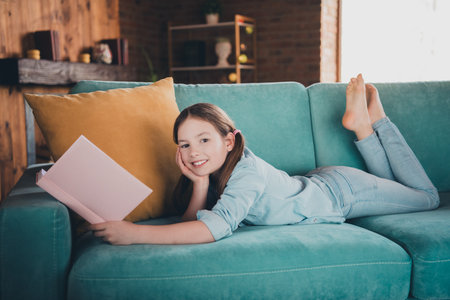 Full length portrait of cute little girl laying sofa read book wear shirt loft interior modern flat indoorsの写真素材