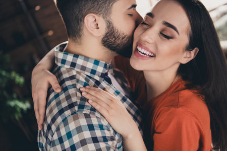 Photo of cheerful cute young couple cuddle hugging kiss cheek enjoy weekend have fun buy new apartment indoors inside house homeの写真素材