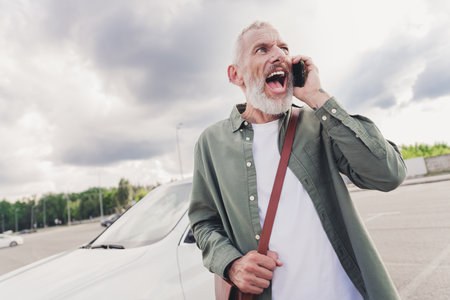 Photo of furious angry moody guy dressed khaki shirt vehicle crash screaming modern device outdoors urban city streetの写真素材