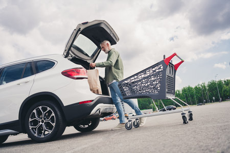 Full body photo of senior man putting paper bags food car trunk shopping cart mall parking