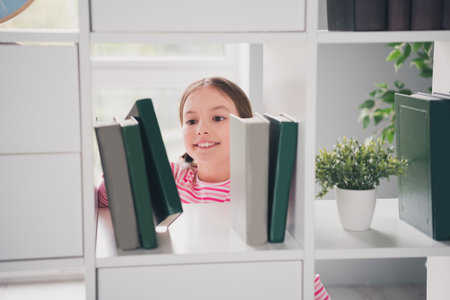 Photo portrait of cute small schoolgirl peek books library sheld interested home education school interiorの写真素材