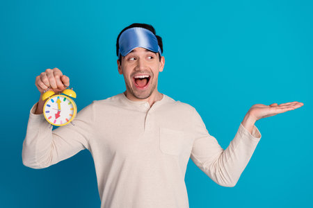 Portrait of nice young man hold clock empty space wear pajama isolated on blue color backgroundの写真素材