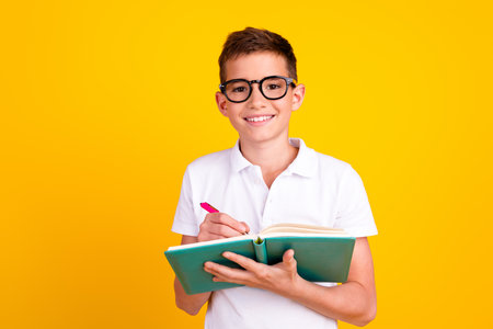 Photo of cheerful positive boy dressed polo t-shirt writing diary emtpy space isolated yellow color...