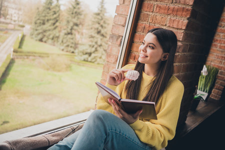 Photo portrait of nice teen lady read book sit windowsill eat snacks dressed yellow garment spend pastime spacious house indoors roomの写真素材