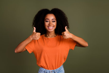 Photo of satisfied woman with perming coiffure dressed orange shirt showing thumbs up approve good job isolated on dark khaki backgroundの写真素材