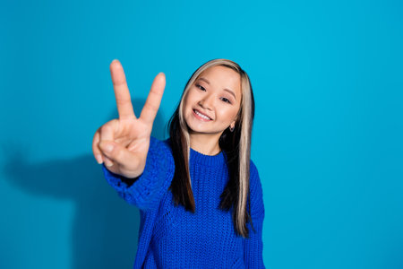 Photo portrait of lovely young lady cheerful v-sign dressed stylish knitwear garment isolated on blue color backgroundの写真素材