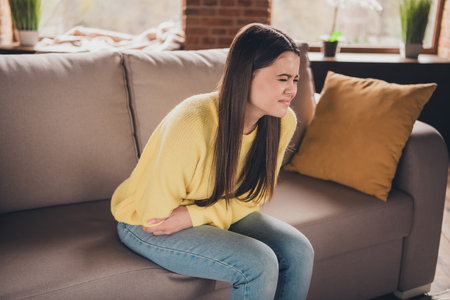 Photo portrait of attractive teen woman sit sofa suffer stomach pain dressed yellow outfit cozy day light home interior living roomの写真素材