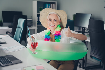 Photo of boss mature woman thumb up ready summer vacation swimming ring dressed formalwear comfortable light startup office room interiorの写真素材