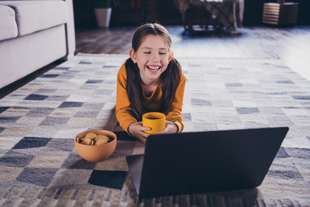 Full body photo of nice little girl lying floor netbook watch cartoon snacks drink tea wear orange...