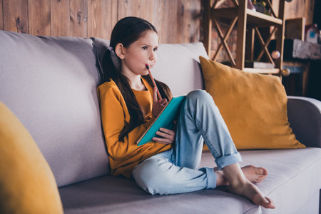 Full body photo of nice little girl sit couch minded write homework wear orange clothes modern interior flat indoorsの写真素材
