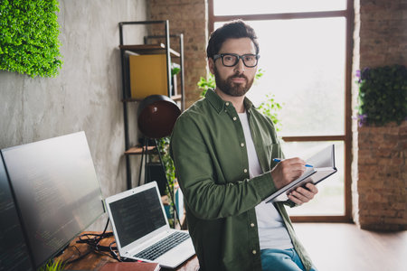 Portrait of professional hacker young man notepad pen computer desk loft interior office indoorsの写真素材