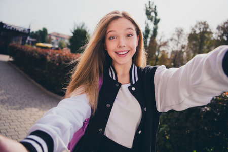 Photo of excited good mood girl dressed bomber walking college recording video vlog outdoors urban...