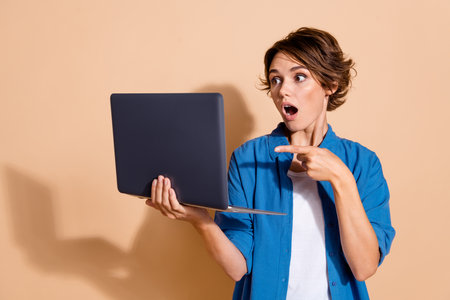 Photo of young funny working woman crm manager pointing finger her laptop with data issues shocked isolated over beige color backgroundの写真素材