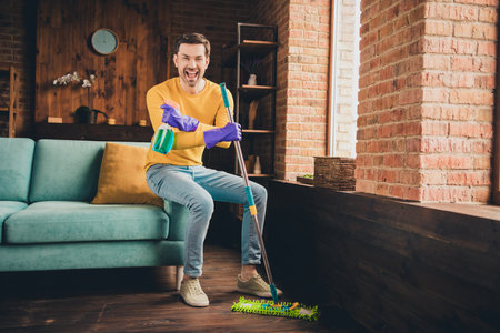 Full length photo of young guy mop floor clean house sit couch excited hold detergent dressed yellow...