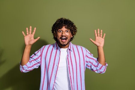 Portrait photo of handsome arabian businessman in striped casual shirt and eyeglass raised hands up amazed isolated on khaki color backgroundの写真素材