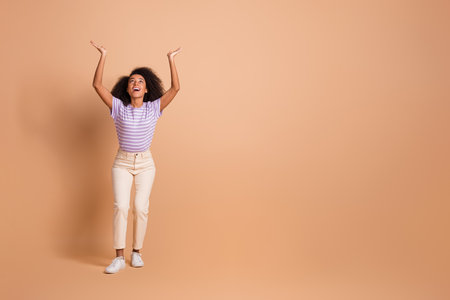 Full length portrait of pretty young woman look up arms hold empty space wear t-shirt isolated on beige color backgroundの写真素材