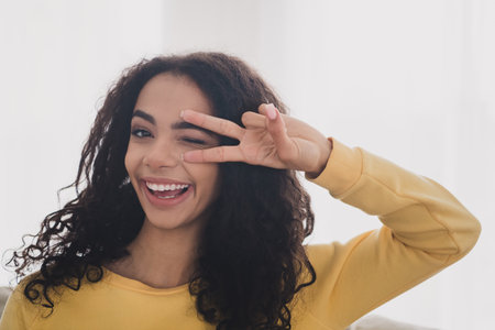 Photo portrait of wavy black haired model young girl wearing yellow shirt blink eye cover face v sign isolated indoors living room with daylightの写真素材