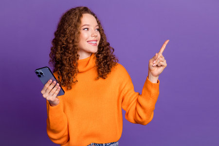 Photo of lovely young girl hold phone direct finger empty space wear orange pullover isolated on violet color backgroundの写真素材