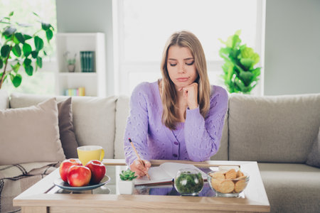 Photo of lovely cheerful girl sit couch table write notebook moody unsure dressed violet garment spend pastime spacious house indoors roomの写真素材