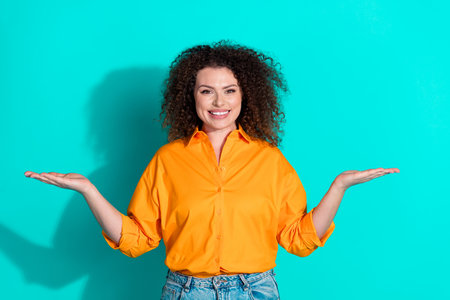 Photo of positive nice woman with wavy hair dressed orange t-shirt palms comparing objects empty space isolated on teal color backgroundの写真素材