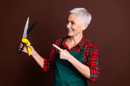 Photo of positive short grey hair woman gardener wear red checkered shirt hold pliers isolated on brown color backgroundの写真素材