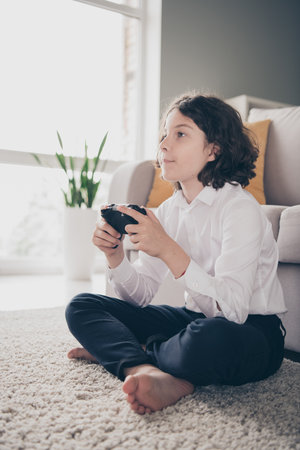 Photo of cute cheerful little boy schoolkid sitting floor playing game home apartment room interior...