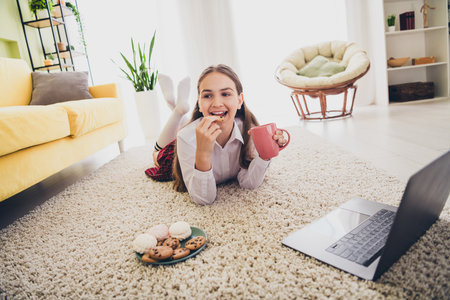 Full length photo of adorable teen girl watch movie drink tea eat snacks lying floor wear uniform remote lesson education from home indoorsの写真素材