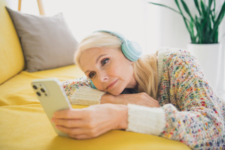 Portrait of nice aged lady headphones use phone wear sweater living room flat indoorsの写真素材