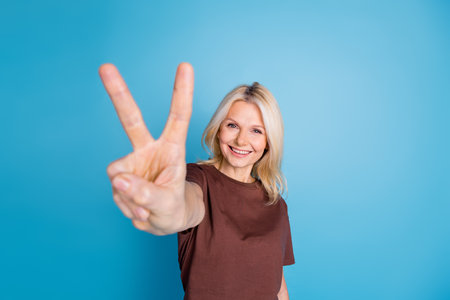 Photo of positive nice lovely senior woman wear trendy brown clothes demonstrate v-sign isolated on blue color backgroundの写真素材