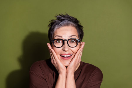 Photo portrait of gray hair beautiful grandmother in brown t shirt and eyeglasses touch cheeks amazing news isolated on khaki color backgroundの写真素材