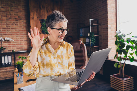 Photo of attractive retired woman netbook wave hand video call wear formalwear comfortable modern office room interior indoors workspaceの写真素材
