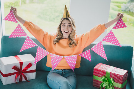Photo of attractive young woman sit sofa excited celebrate birthday hold flags dressed orange outfit...