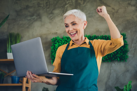 Photo of good mood lucky senior woman dressed apron nursing flowers getting likes device indoors house workplaceの写真素材