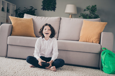 Photo of charming cheerful little boy schoolkid sitting floor playing game home apartment room interior indoorsの写真素材