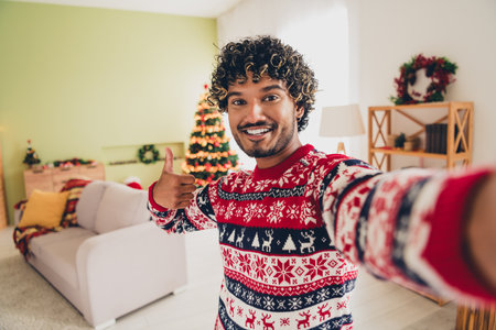 Portrait of nice young american man show thumb up make selfie wear xmas sweater holiday atmosphere flat indoorsの写真素材