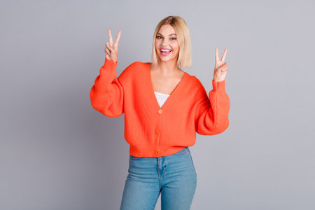 Photo of pretty young girl demonstrate v-sign orange cardigan isolated on grey color background