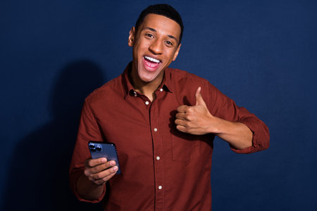 Photo of funky cool guy dressed brown shirt chatting modern device showing thumb up isolated blue...