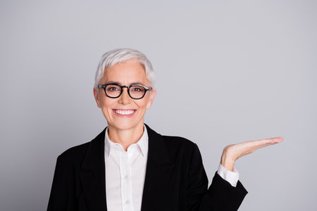 Photo portrait of pretty senior female professional entrepreneur hold empty space wear formalwear specs isolated on gray color backgroundの写真素材