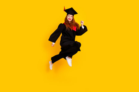 Photo of cheerful positive girl college student wear black robe jump up isolated on yellow color backgroundの写真素材